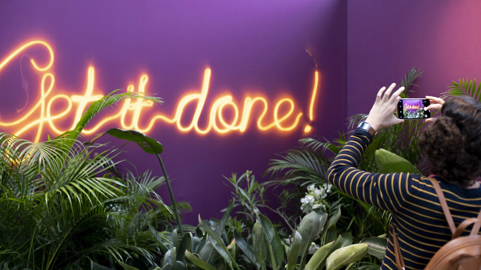 Enseigne lumineuse « Get it done! » sur un mur violet entouré de plantes.