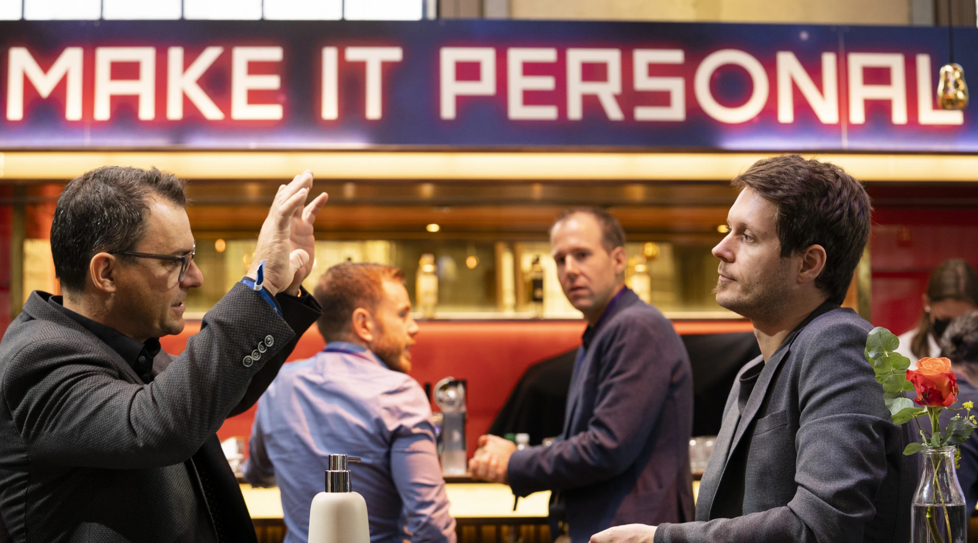 Participants discutant au bar sous la typographie lumineuse « Make it personal ».